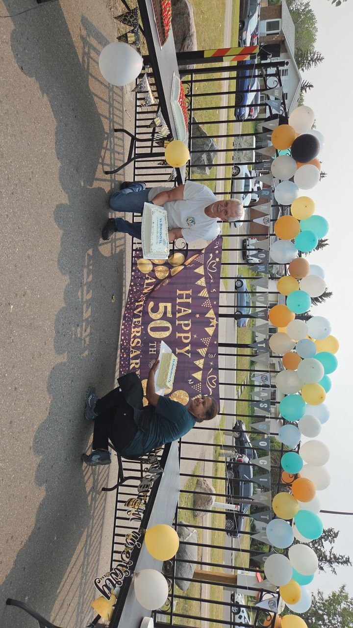 Two volunteers holding the Whitehorn 50th anniversary cakes in front of a Happy 50th Anniversary banner and balloon arch